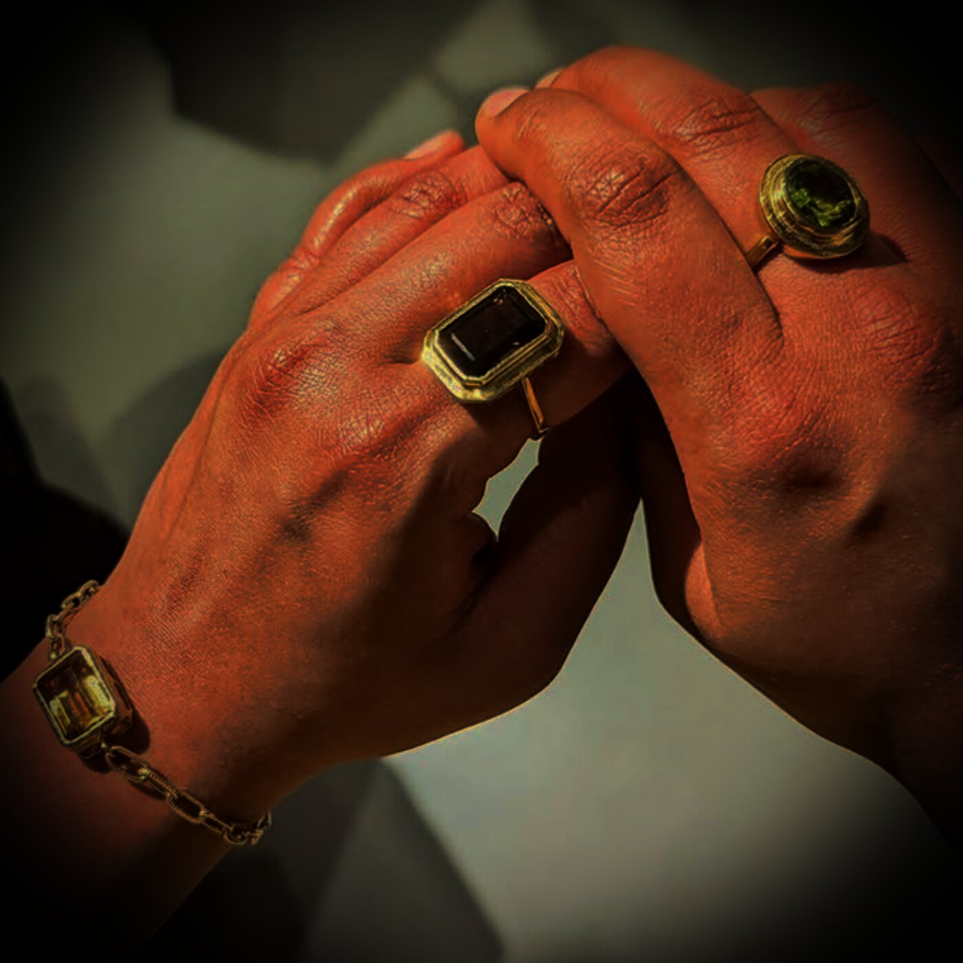 Two clasped hands wearing a smoky quartz ring, a peridot ring and a citrine bracelet by Mor and Mogra, lit by warm gold light.