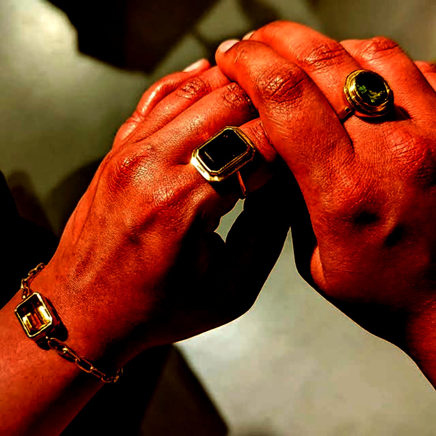 Two hands clasped, wearing gold signet rings, a green-stone ring and a citrine bracelet.